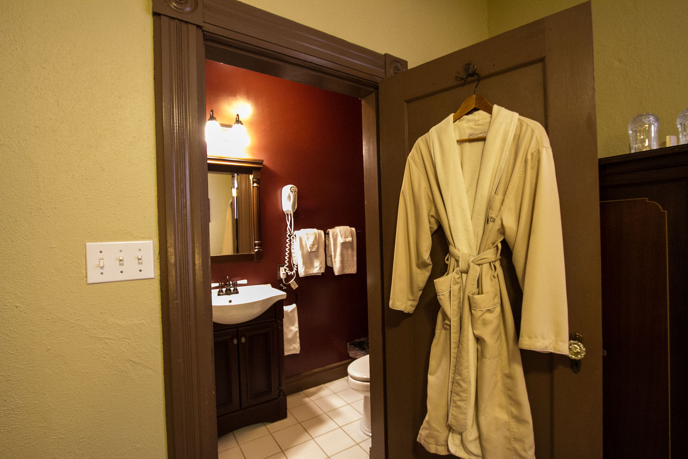 A view from a bedroom into a small, attached private bathroom with deep red walls, featuring a dark wood vanity, white sink, and a plush cream-colored bathrobe hanging on the open door.