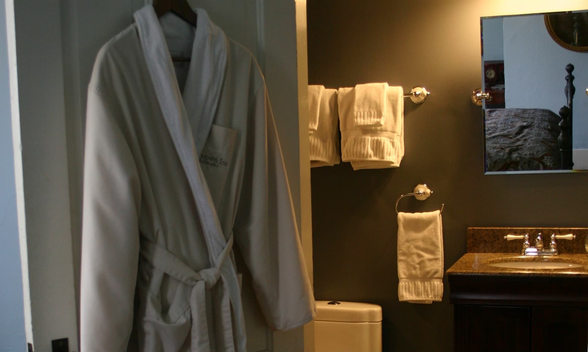 A cream-colored, plush bathrobe hanging on an open white door, leading into a contemporary private bathroom with dark gray walls and a wood vanity.