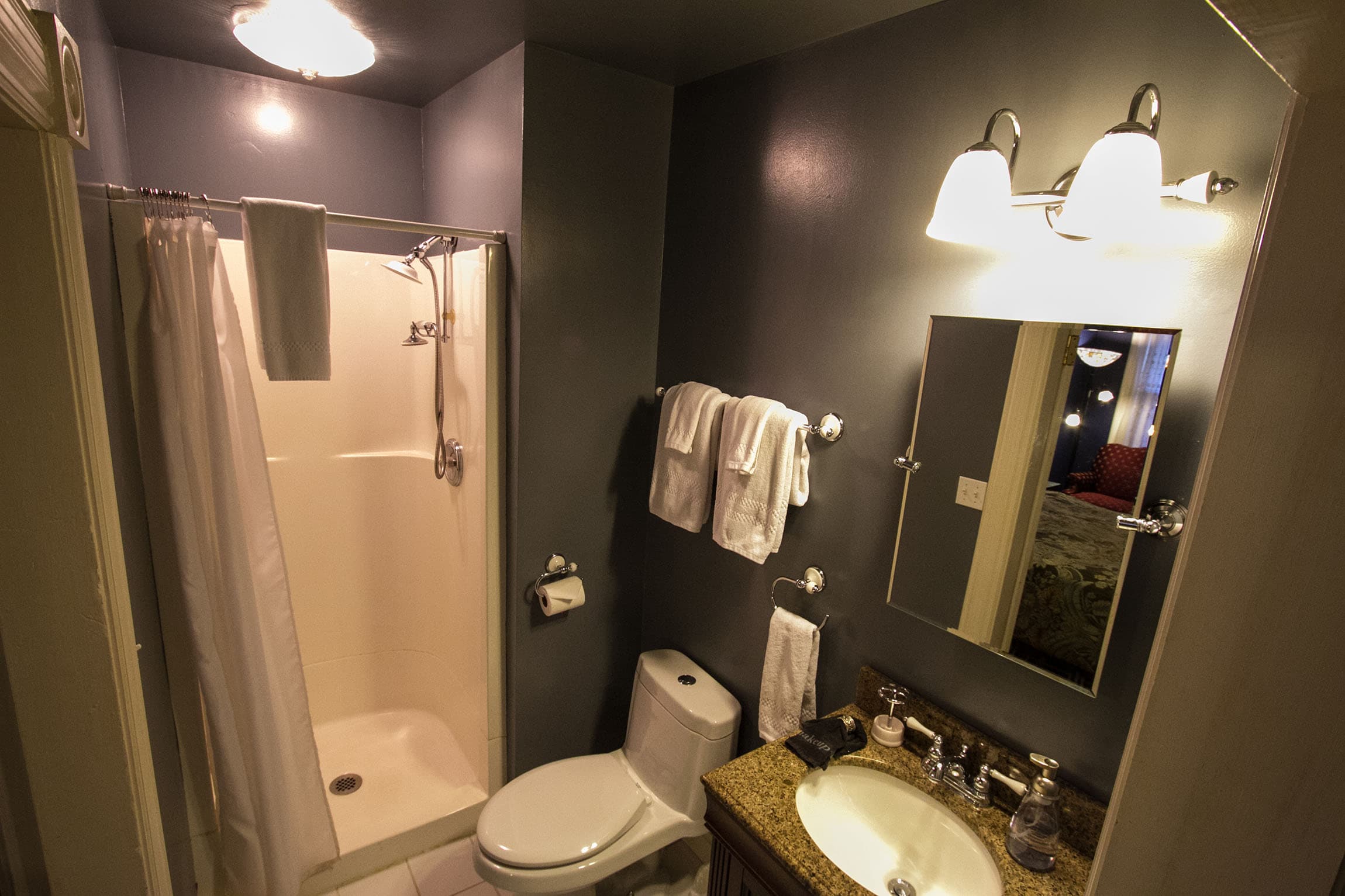 A high-angle view of a modern private bathroom  featuring a white walk-in shower with a curtain, a toilet, and a vanity with a granite countertop and lighted mirror.
