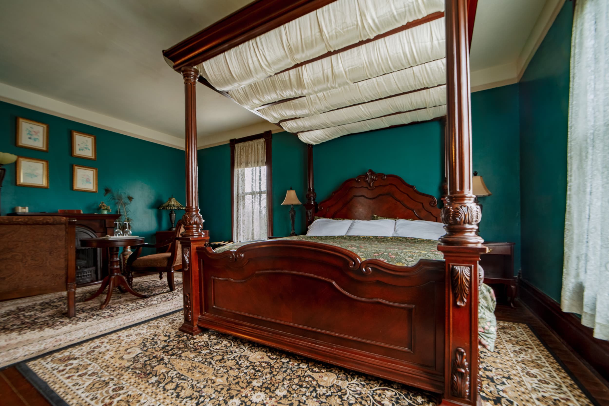 Cozy sitting area featuring a dark wood fireplace, an antique writing desk, and a small lace-covered table set with wine glasses in front of two green armchairs.