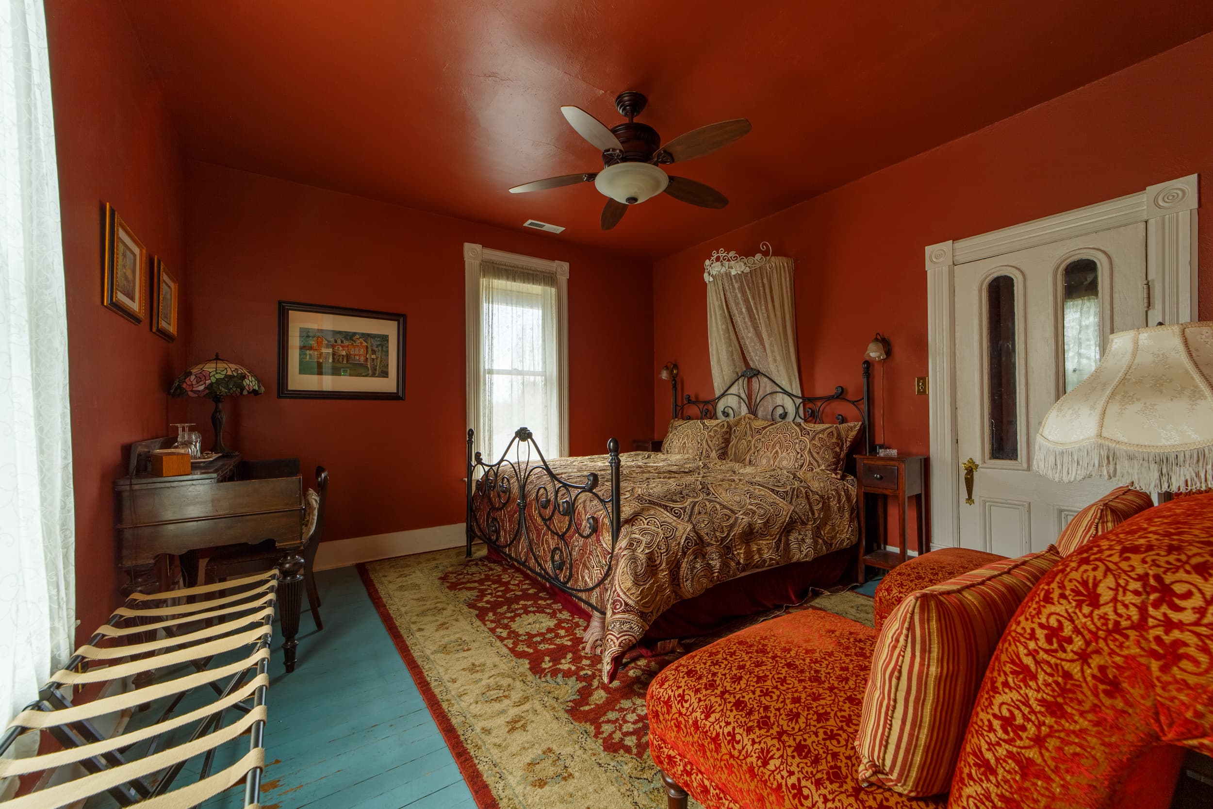 A cozy guest room with deep red walls, featuring a wrought iron king bed with a patterned gold and brown bedspread and white pillows.
