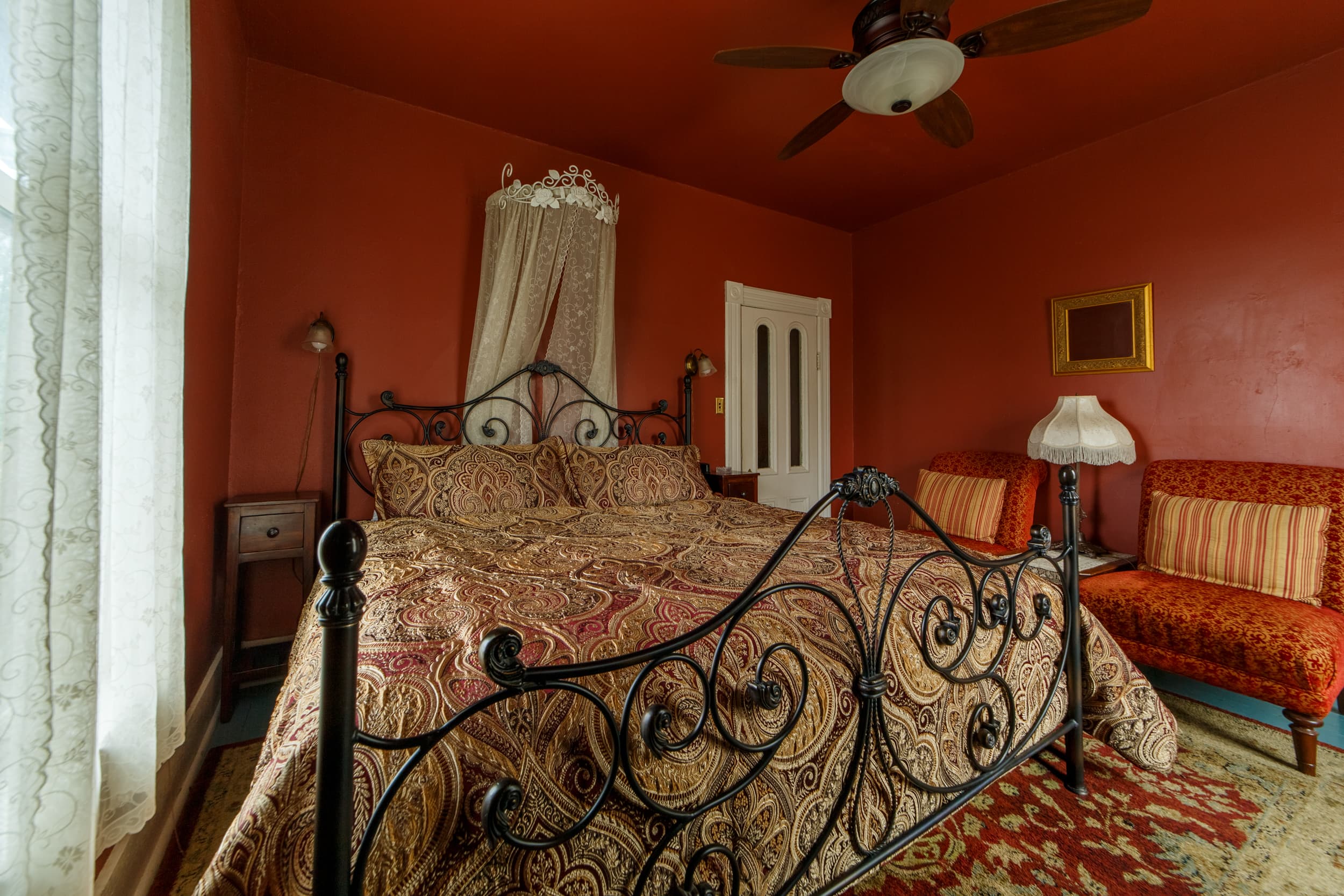 A classic workspace featuring a dark wood writing desk and chair positioned against deep red walls, situated next to a black wrought iron king bed with a gold-patterned bedspread.