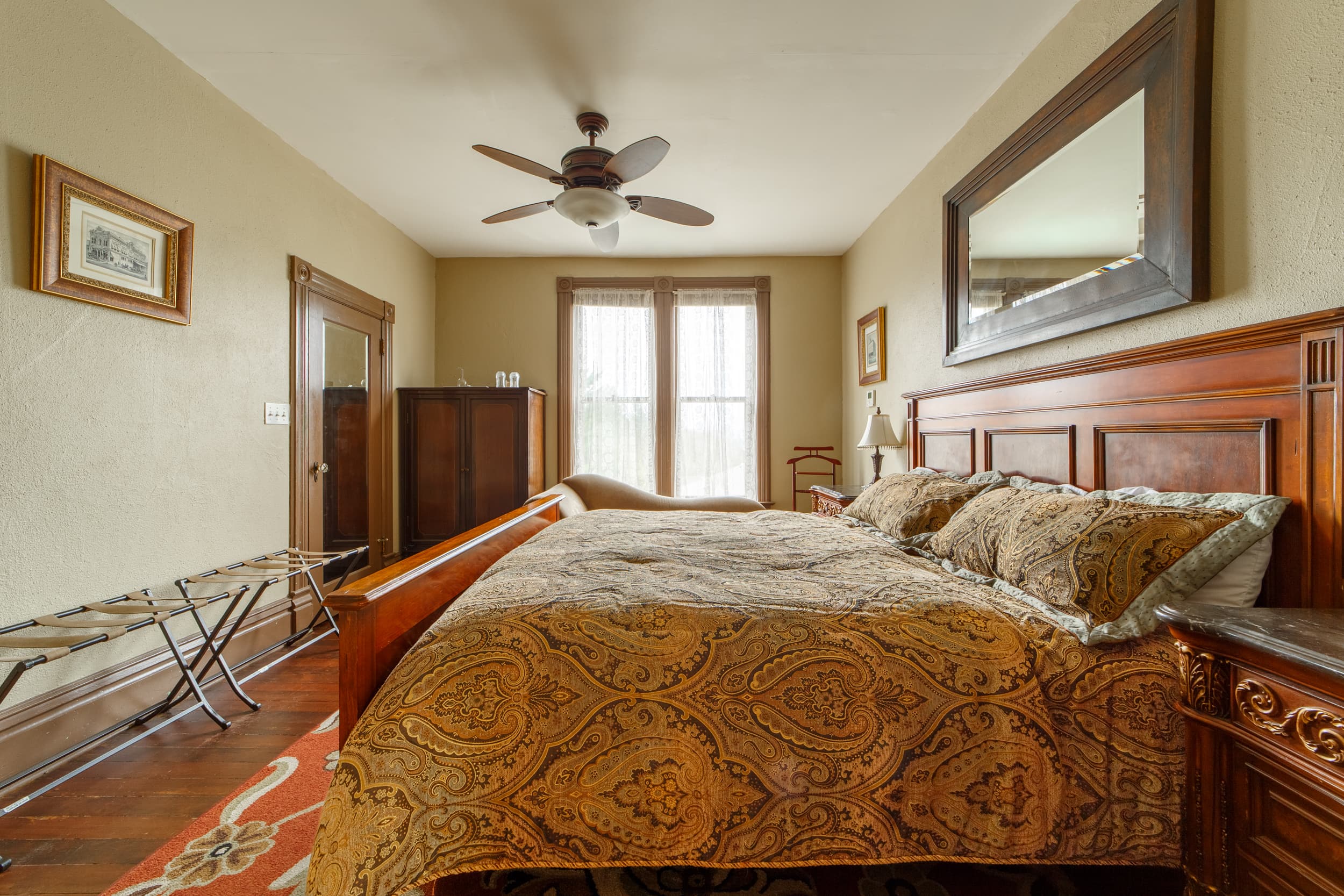 A cozy guest room featuring a large wood-framed king bed with a patterned gold and blue bedspread, a ceiling fan, and a warm bedside lamp.