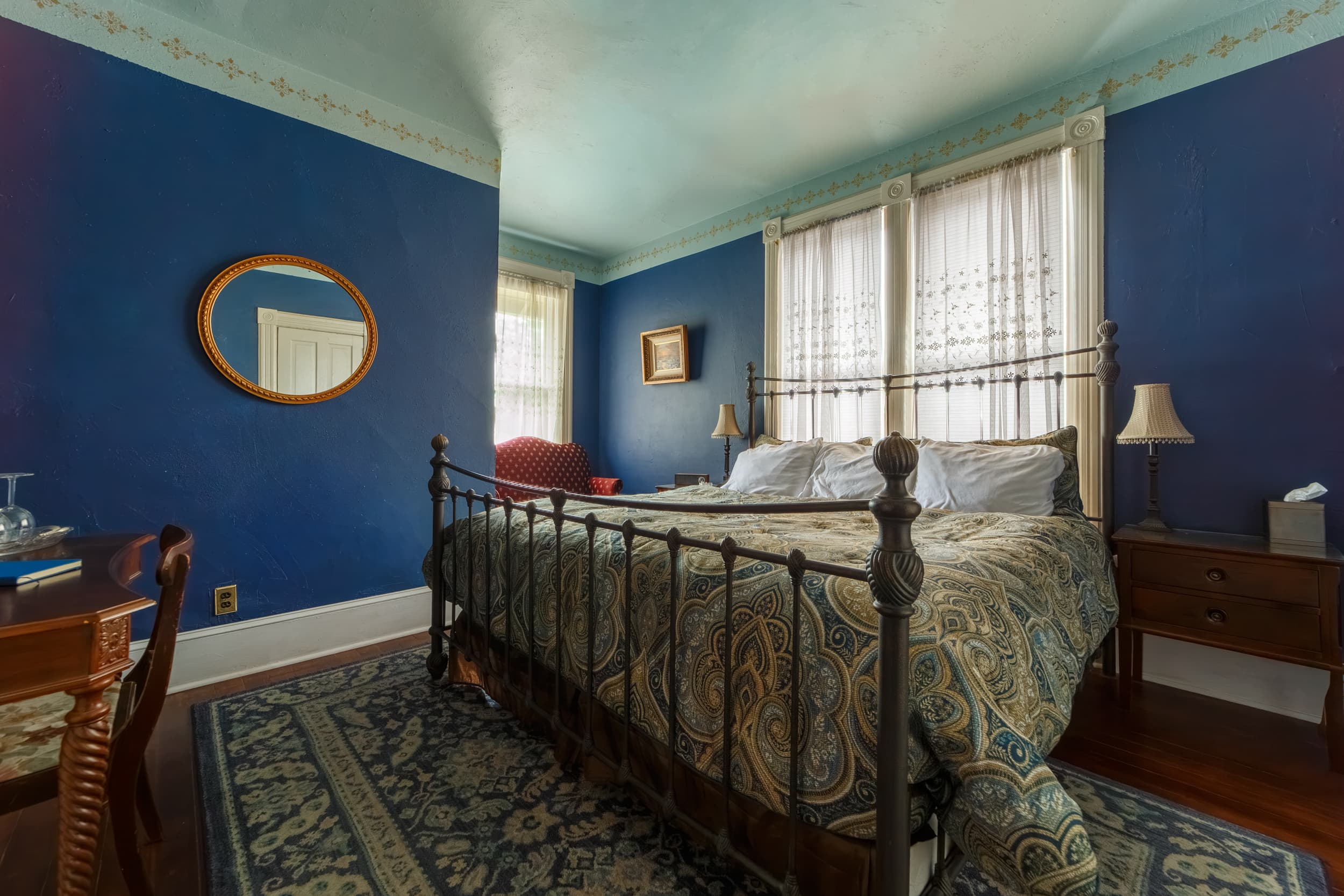 A cozy guest room featuring a wrought iron king bed with a patterned blue and gold bedspread, a red wingback seat in the corner, and a ceiling fan.