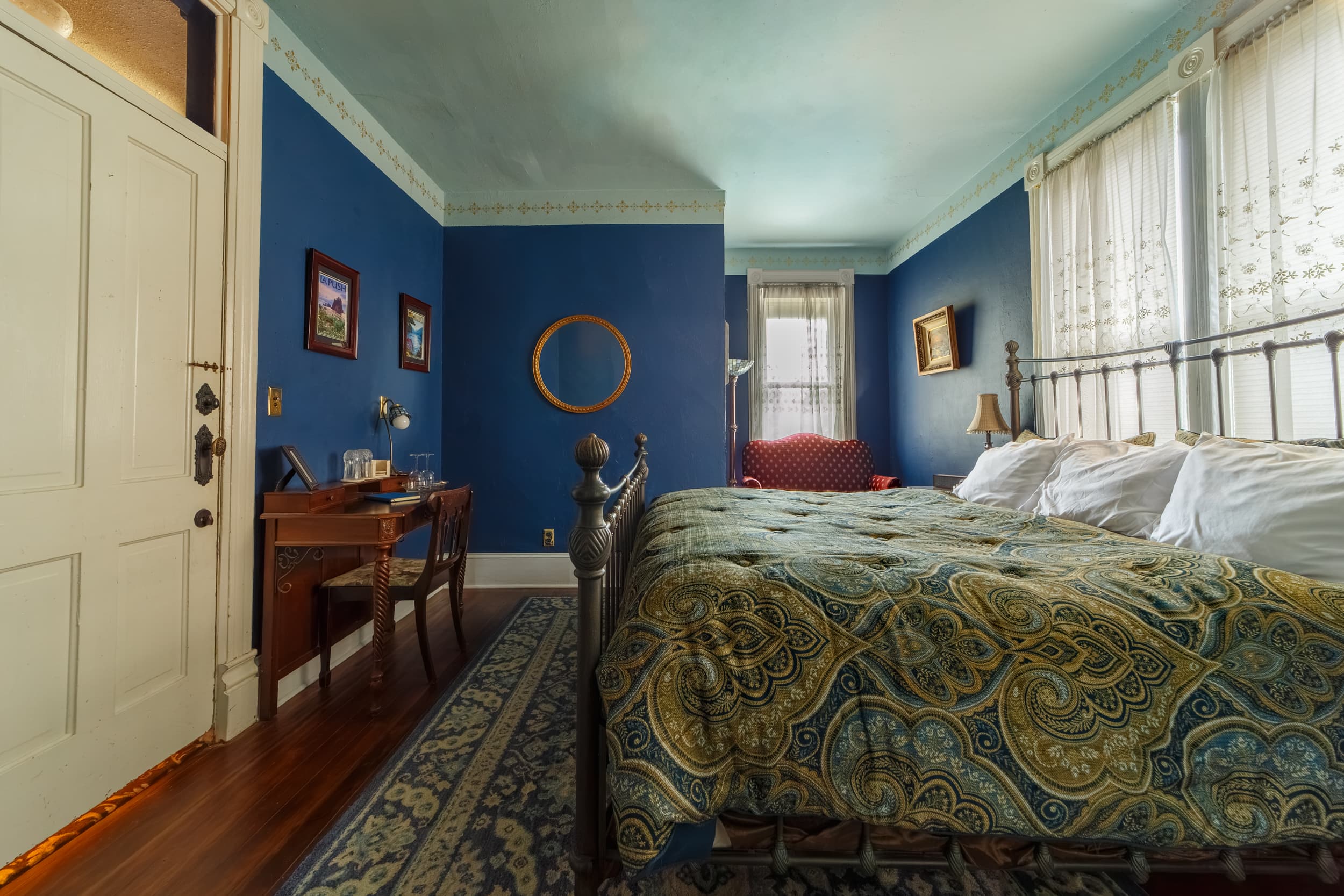 A spacious bedroom with decorative white crown molding, a wrought iron bed with a gold and blue patterned bedspread, and a white door leading to a modern private bathroom where a plush bathrobe is hanging.
