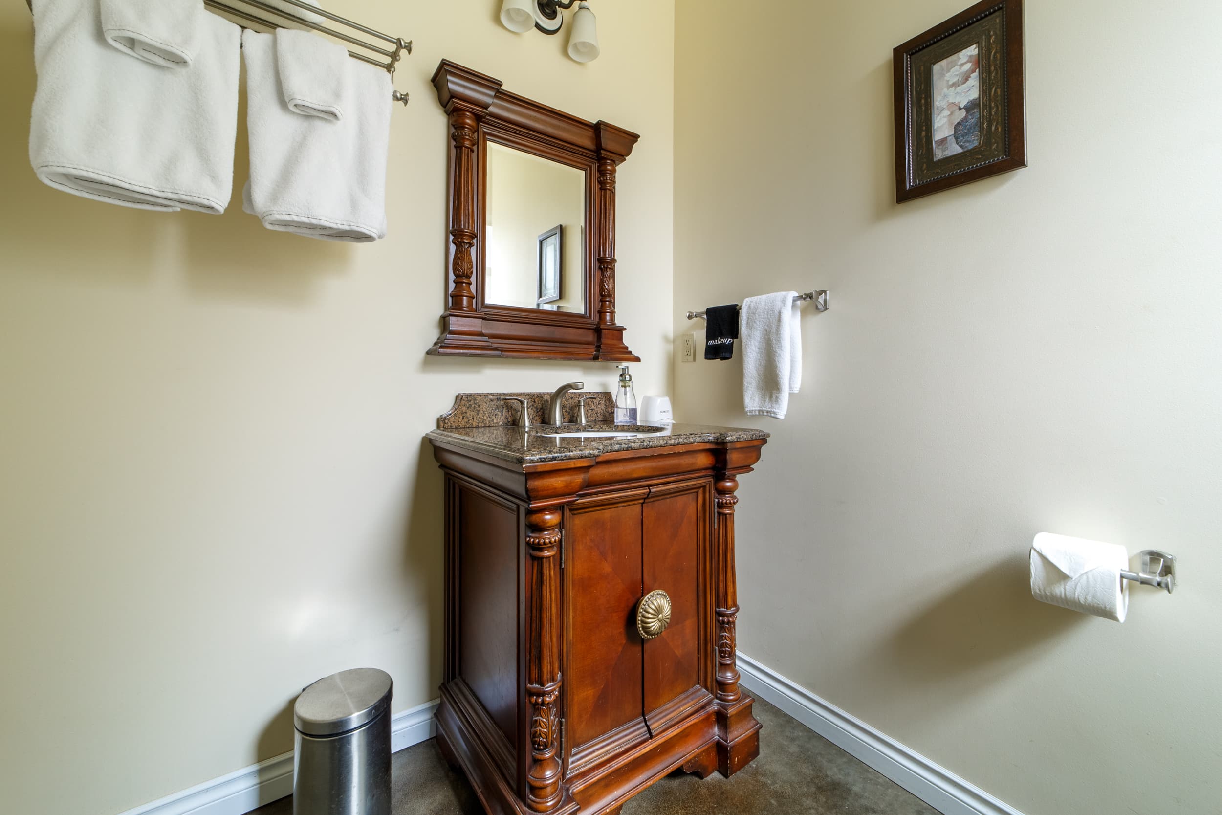 A luxurious, dark wood vanity with an ornate mirror and a granite countertop, featuring classic pillar-style carvings and a gold circular handle on the cabinet door.