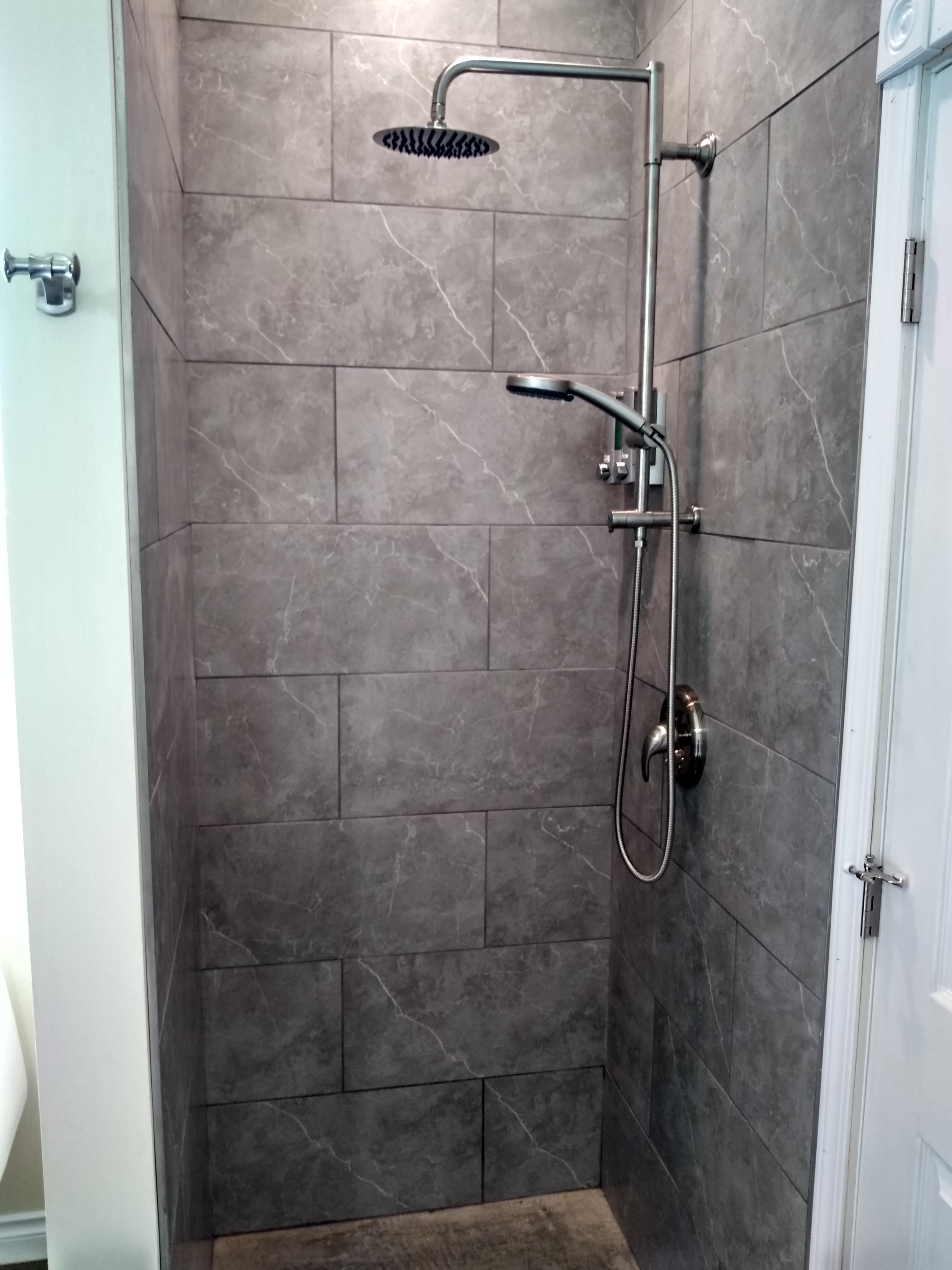 A modern, floor-to-ceiling walk-in shower featuring large, slate-gray stone tiles and a contemporary chrome rain showerhead with an additional handheld sprayer attachment.