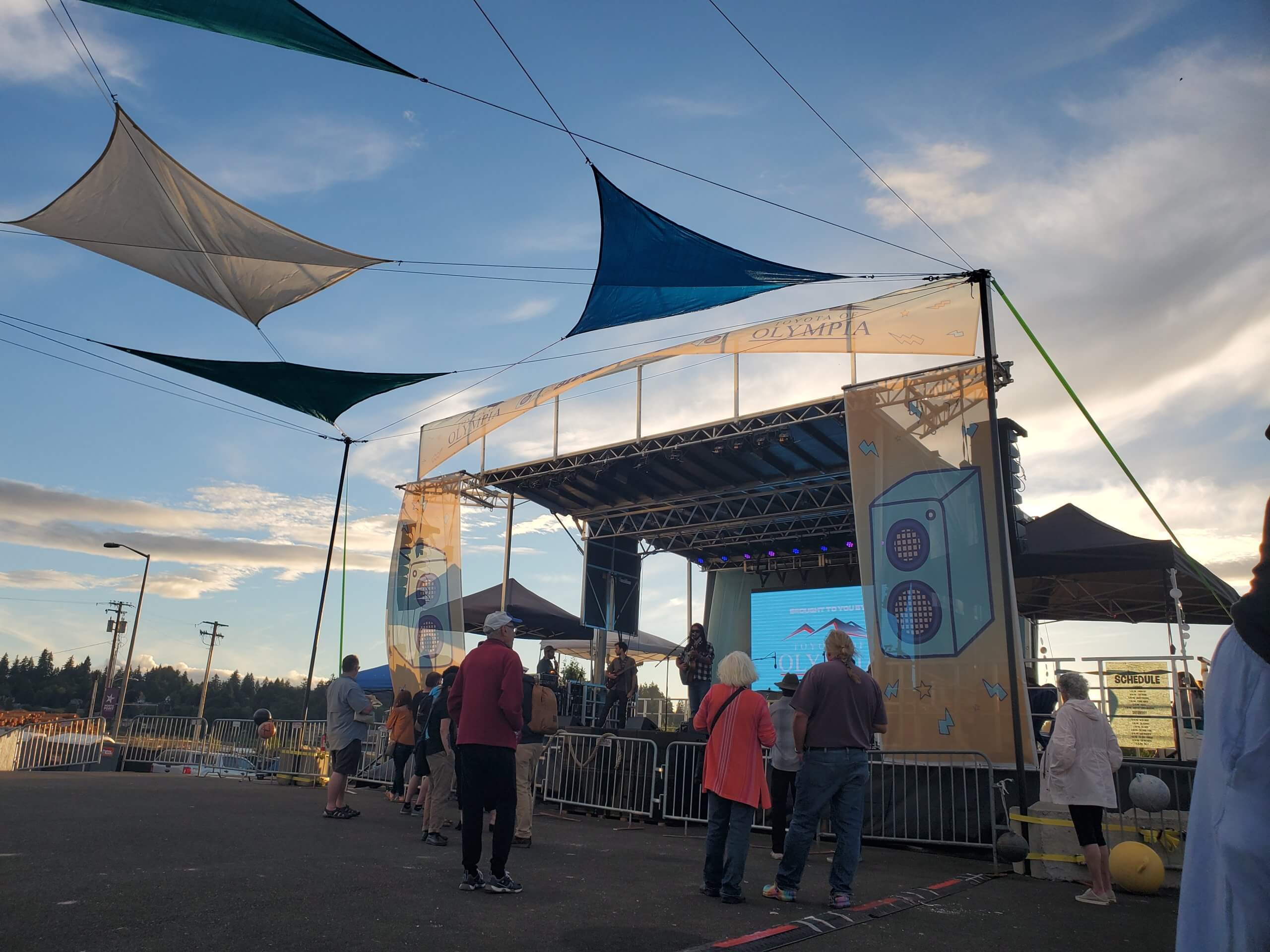 A crowd gathers in front of a stage decorated with colorful sails against a sunset sky.