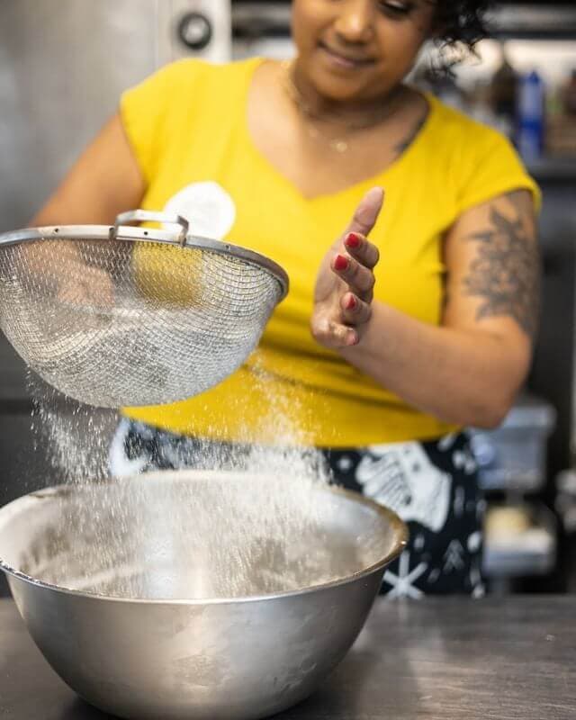A person in a yellow shirt sifts flour into a silver mixing bowl.