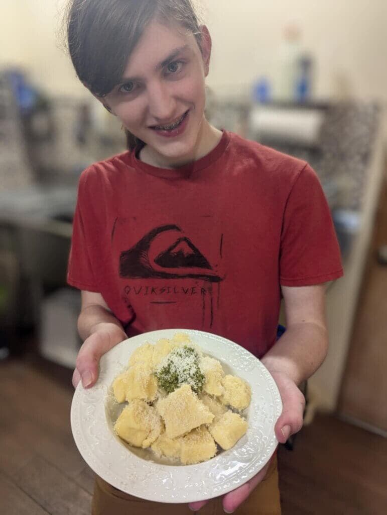 A smiling person in a red shirt holds a plate of pasta topped with cheese and herbs in a kitchen.