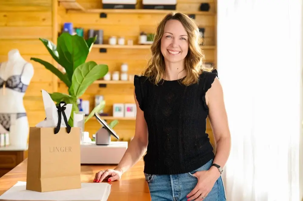 A smiling woman stands behind a counter with a gift bag labeled "LINGER" and greenery in a cozy store setting.