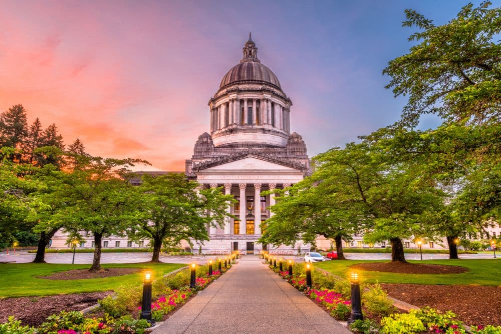 A historic building with a dome is framed by lush trees and flowerbeds under a colorful sunset sky.