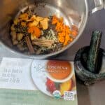 A stainless steel bowl contains a variety of herbs and spices alongside a mortar and a recipe card.