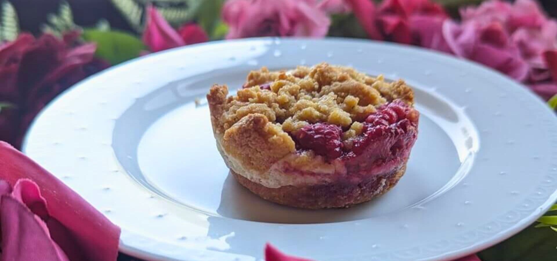 A single raspberry crumble dessert on a white plate, surrounded by pink flowers.