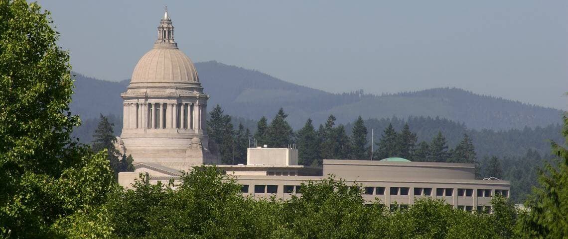 The Washington State Capitol building is framed by trees and mountains in the background.