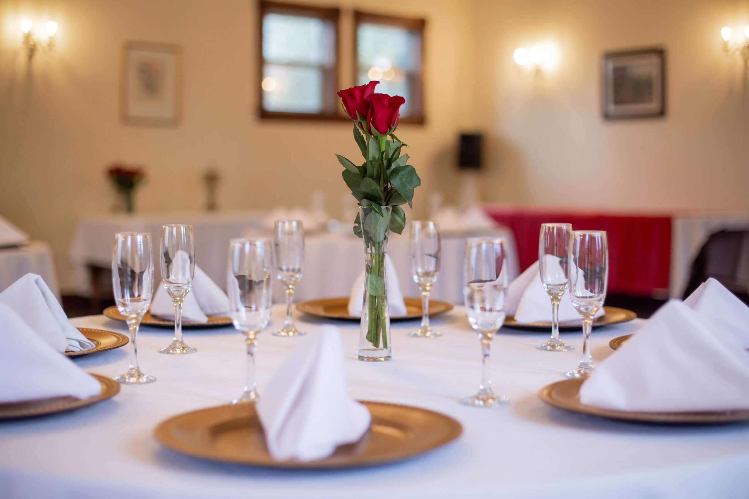 A dining table elegantly set with gold plates, crystal glasses, and a bouquet of red roses in a vase.