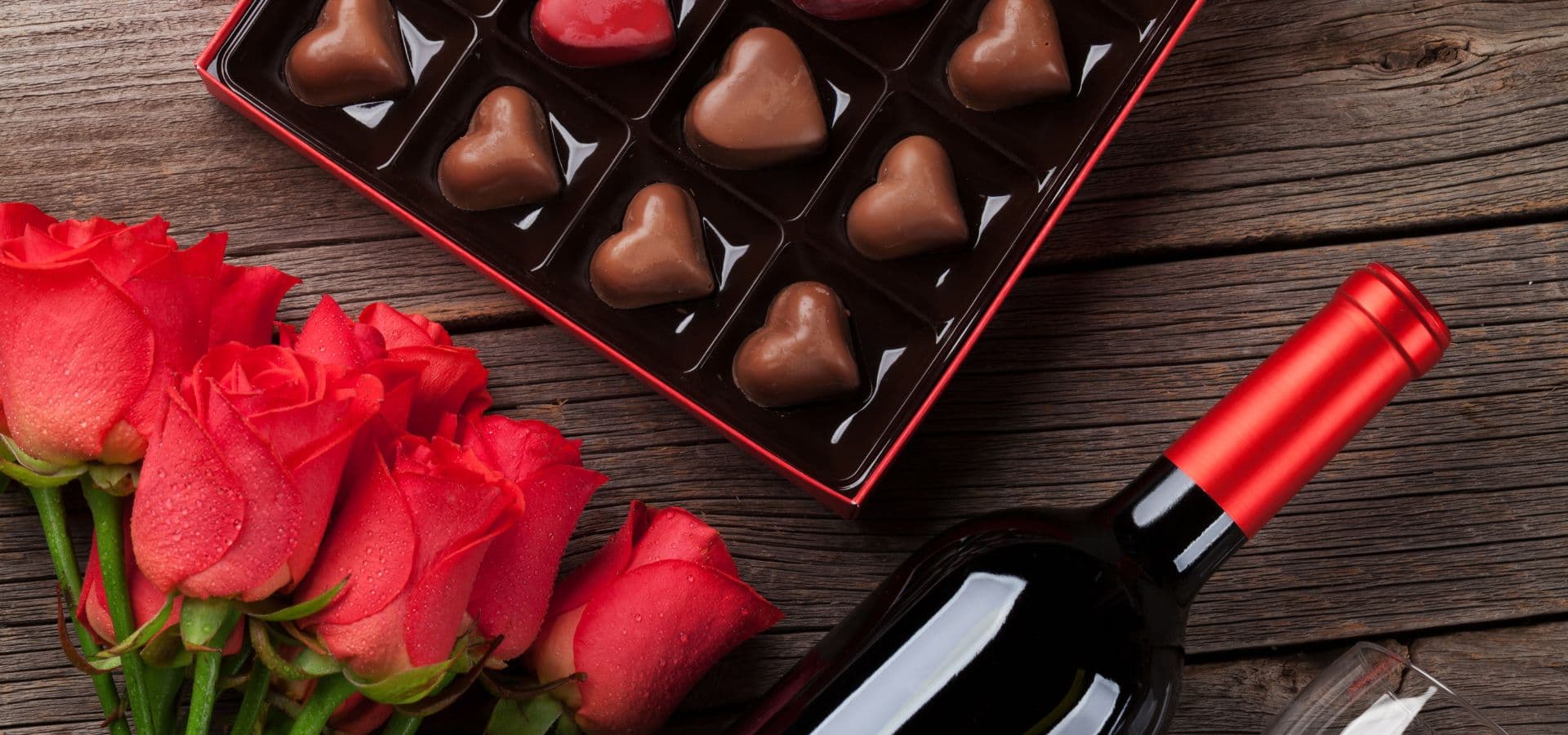 A box of heart-shaped chocolates, a bouquet of red roses, and a bottle of red wine on a wooden surface.