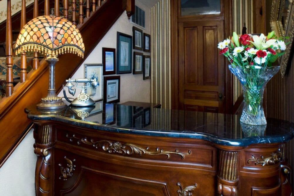 A polished wooden reception desk with a stained glass lamp and a vase of flowers, situated near a staircase and framed certificates on the wall.