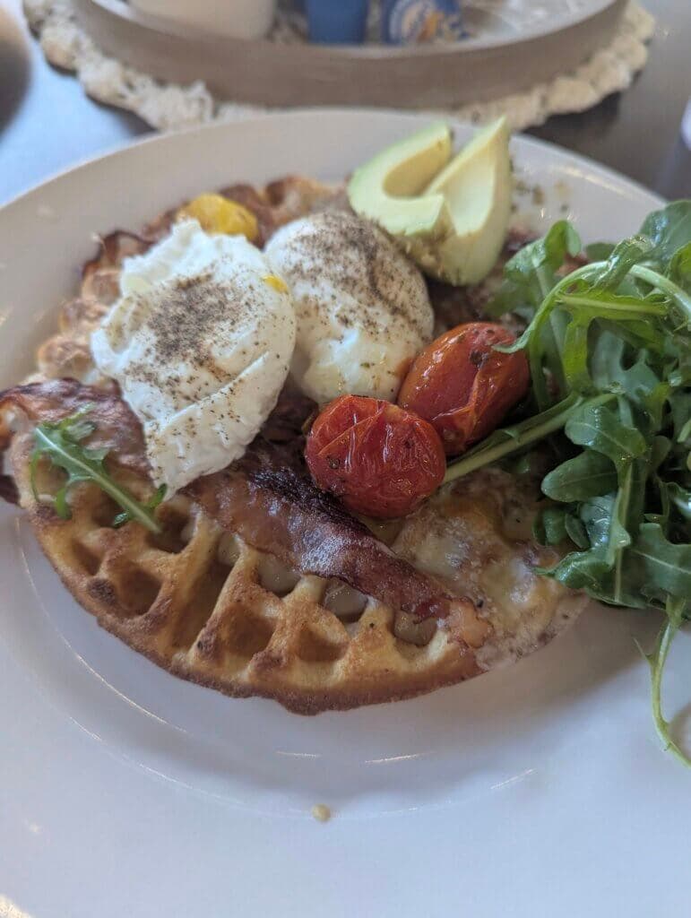 A waffle topped with poached eggs, crispy bacon, roasted cherry tomatoes, avocado slices, and a side of arugula.
