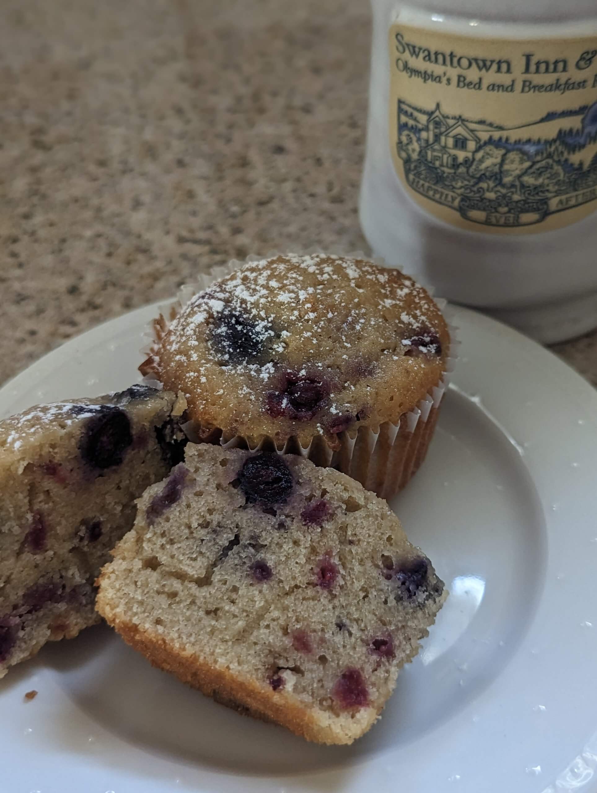 A plate with a muffin topped with powdered sugar and a cut piece revealing berries, alongside a coffee mug.