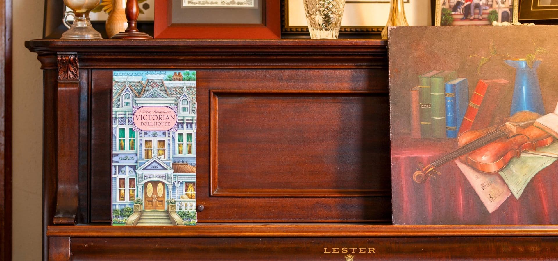 A vintage wooden piano adorned with decorative items, a book about Victorian architecture, and an artwork featuring books and a violin.