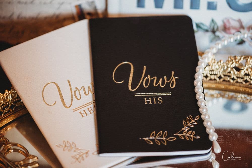 Two vow notebooks, one white labeled "Hers" and one black labeled "His," arranged on a decorative surface with a pearl necklace.