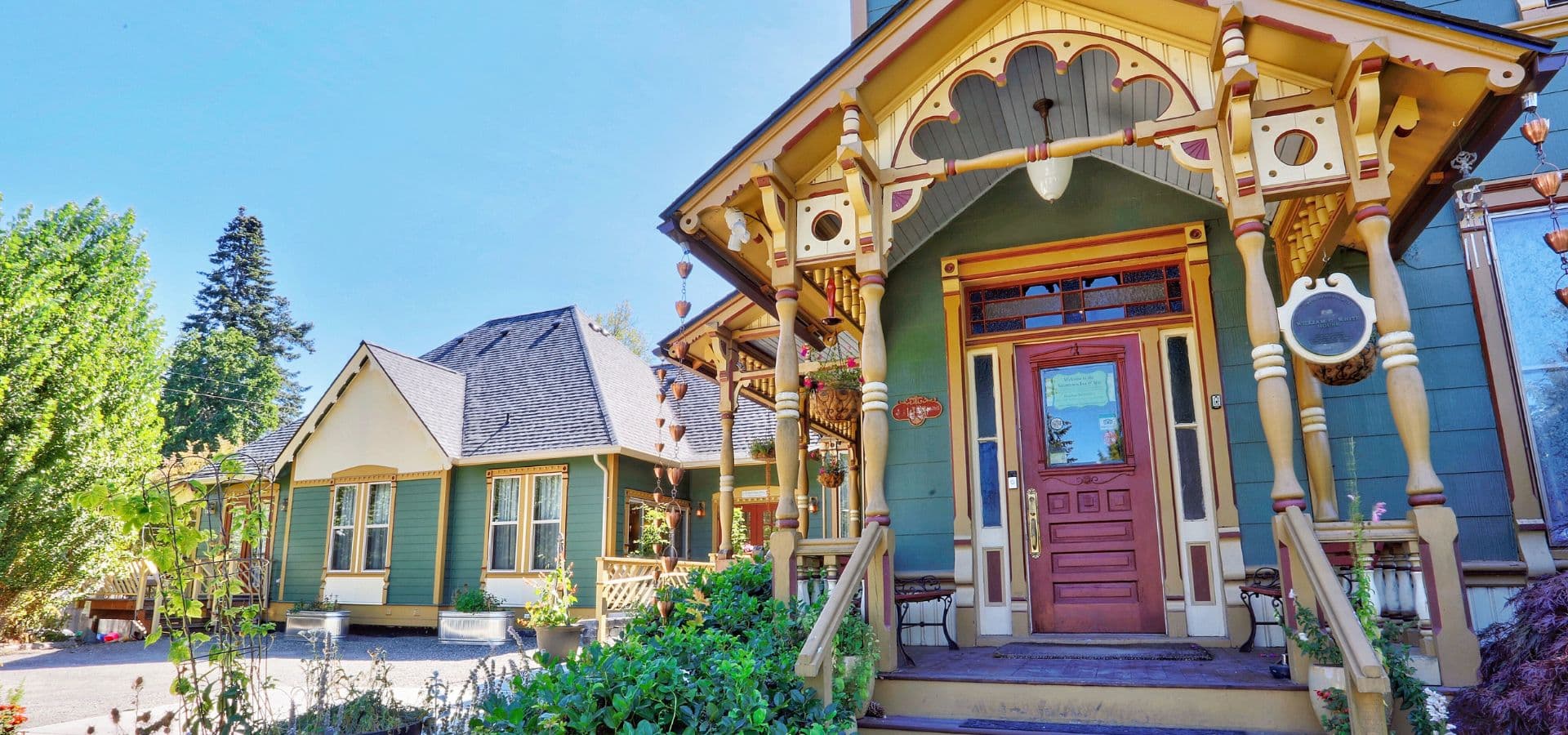 A beautifully detailed, colorful Victorian-style house with ornate porch details and surrounding greenery.