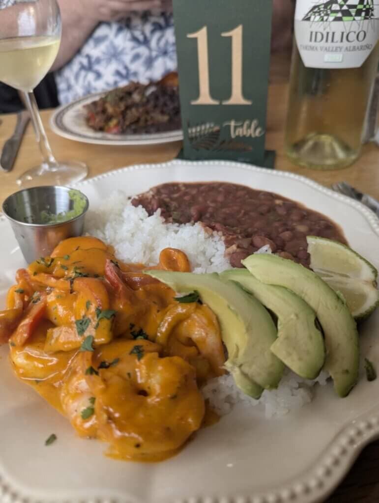 A plate of shrimp in creamy sauce served with rice, beans, sliced avocado, and a small cup of green sauce.