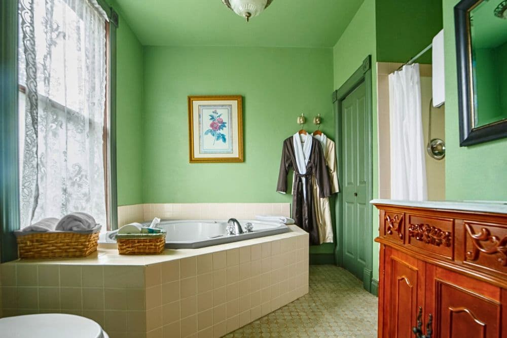 A bright green bathroom featuring a bathtub, towels, two bathrobes hanging, and wooden cabinet details.