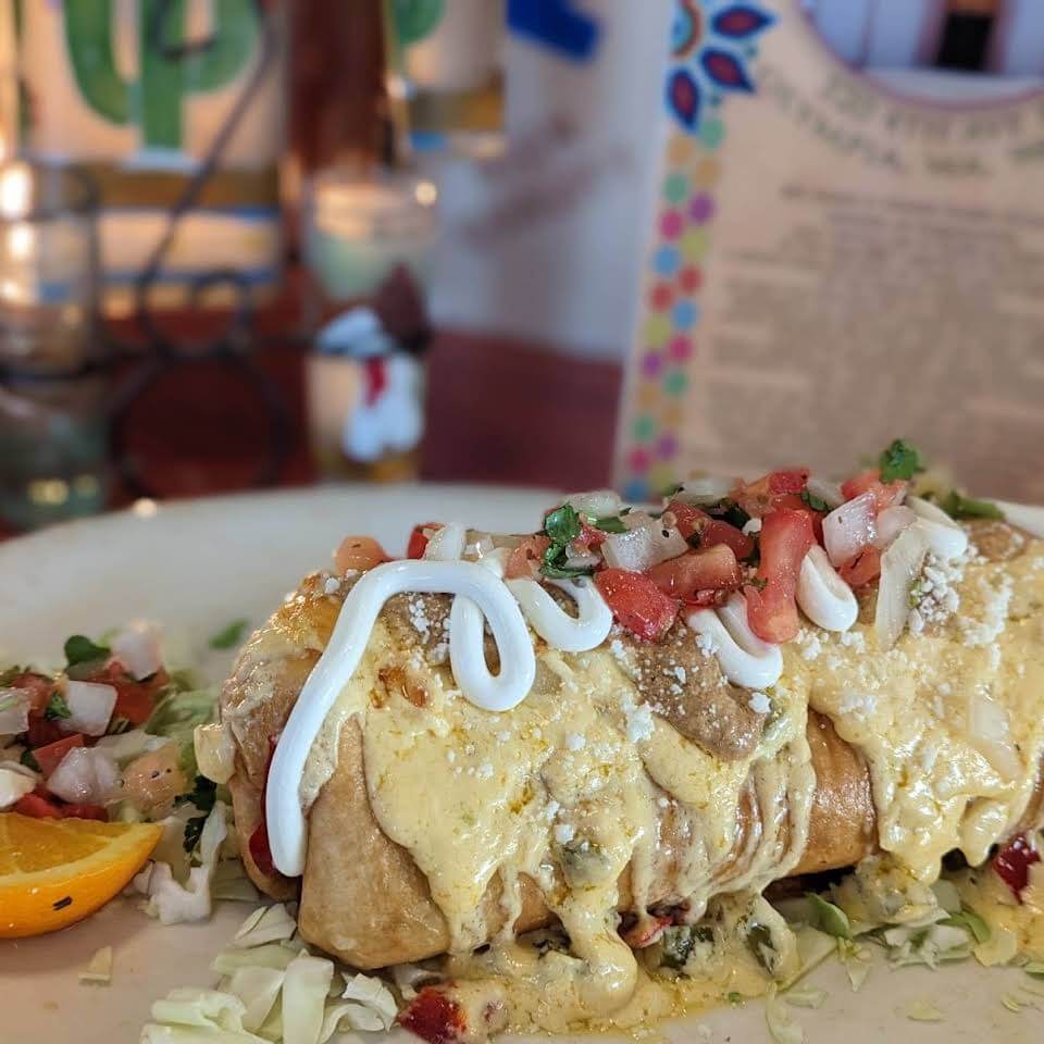 A large burrito topped with creamy sauce, pico de gallo, and sour cream, served on a plate with cabbage and an orange slice.