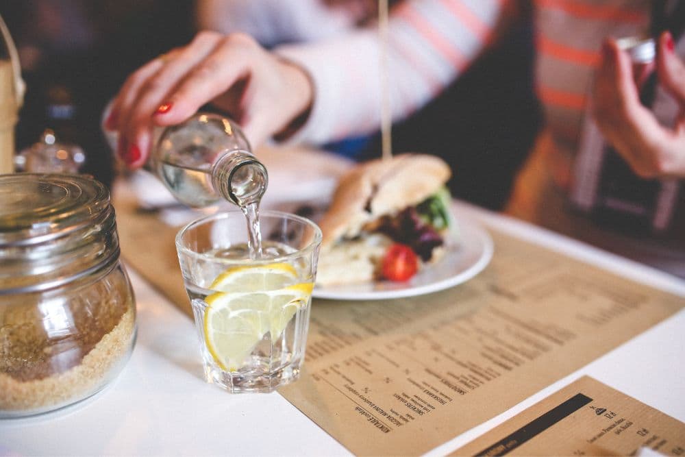A person pours sparkling water into a glass with a lemon slice, accompanied by a sandwich and a menu on the table.
