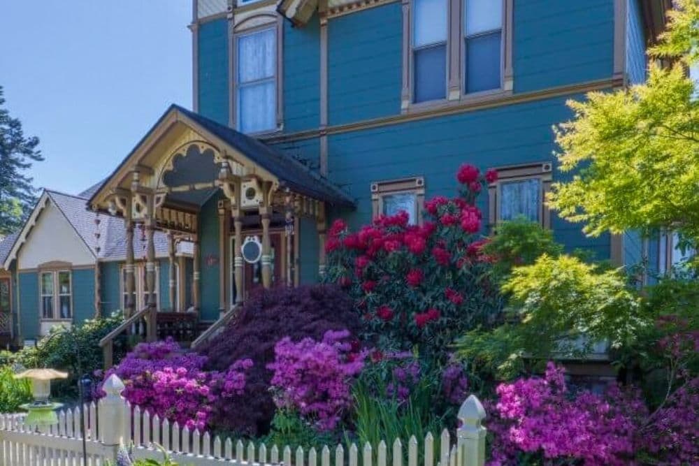 A Victorian house with a teal exterior surrounded by vibrant flowers and a white picket fence.