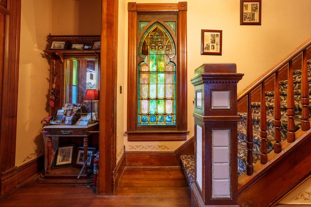 A stained glass window illuminated by sunlight next to a wooden staircase and antique furnishings.