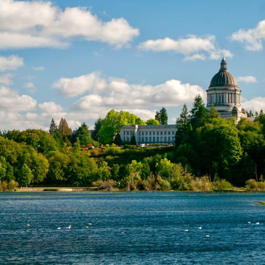 A view of a dome-topped building surrounded by lush trees and a calm lake under a partly cloudy sky.