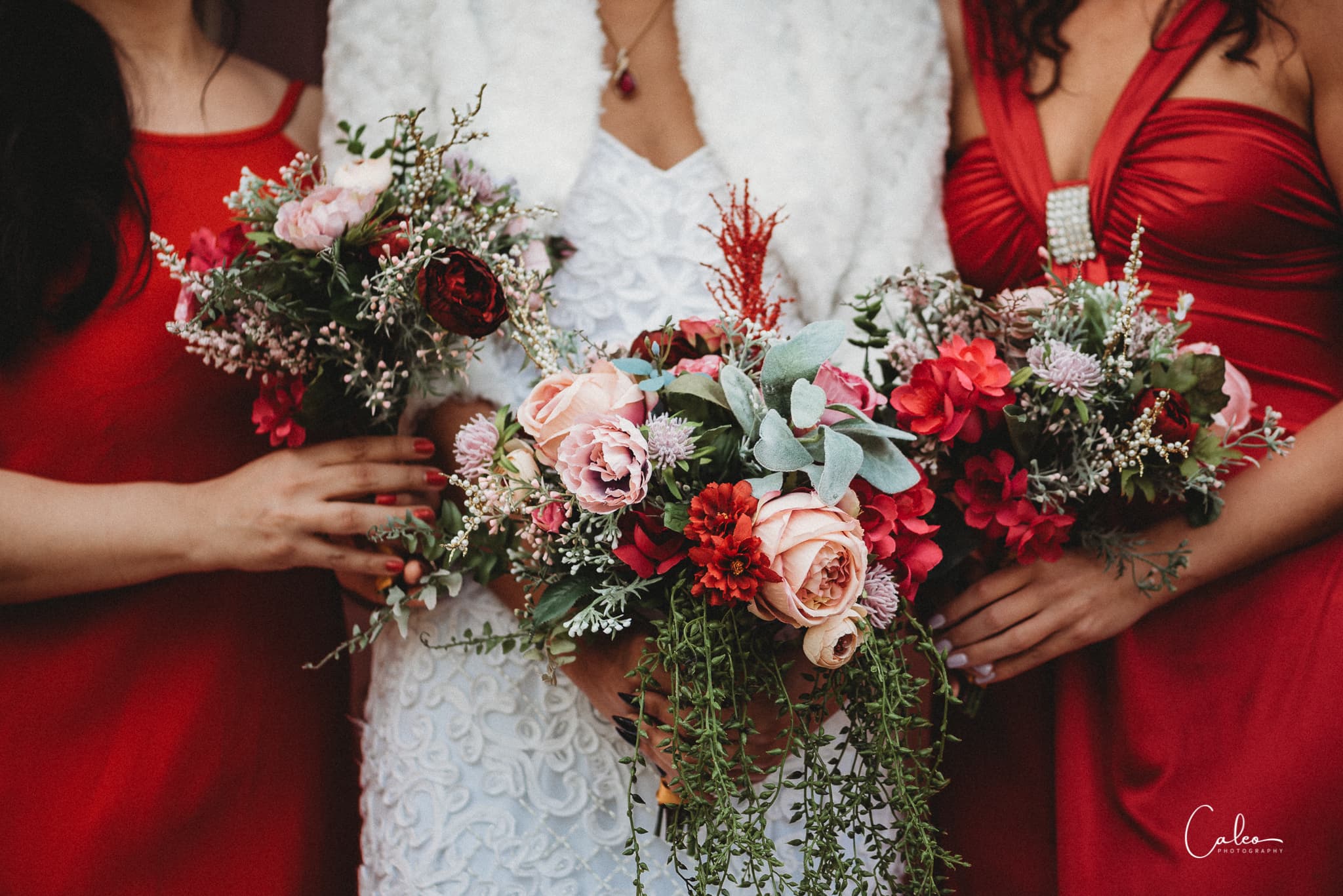 Three women in red dresses hold colorful floral bouquets, with one wearing a white dress and fur shawl.