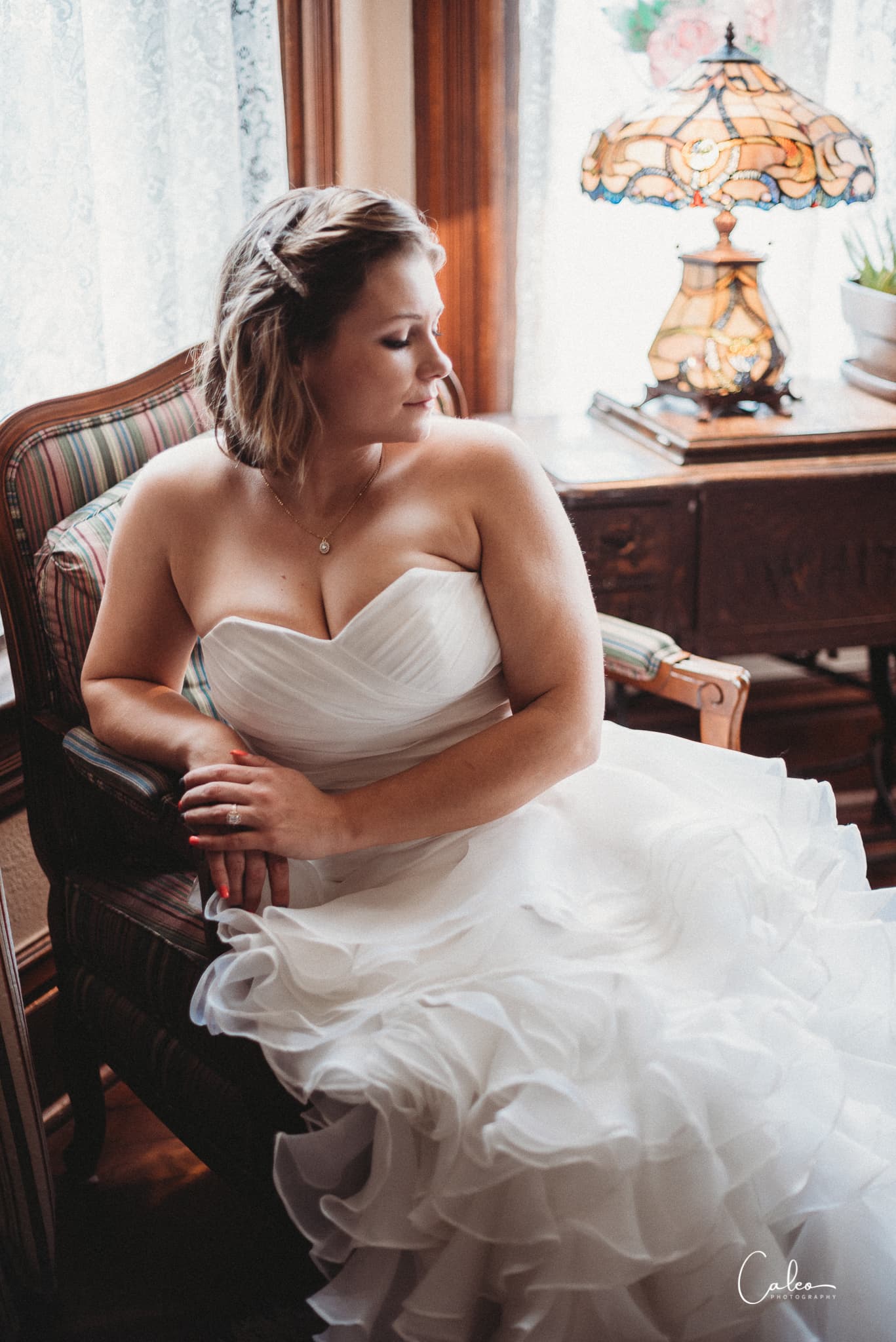 A bride in a white wedding gown sits thoughtfully in a chair beside a vintage lamp.