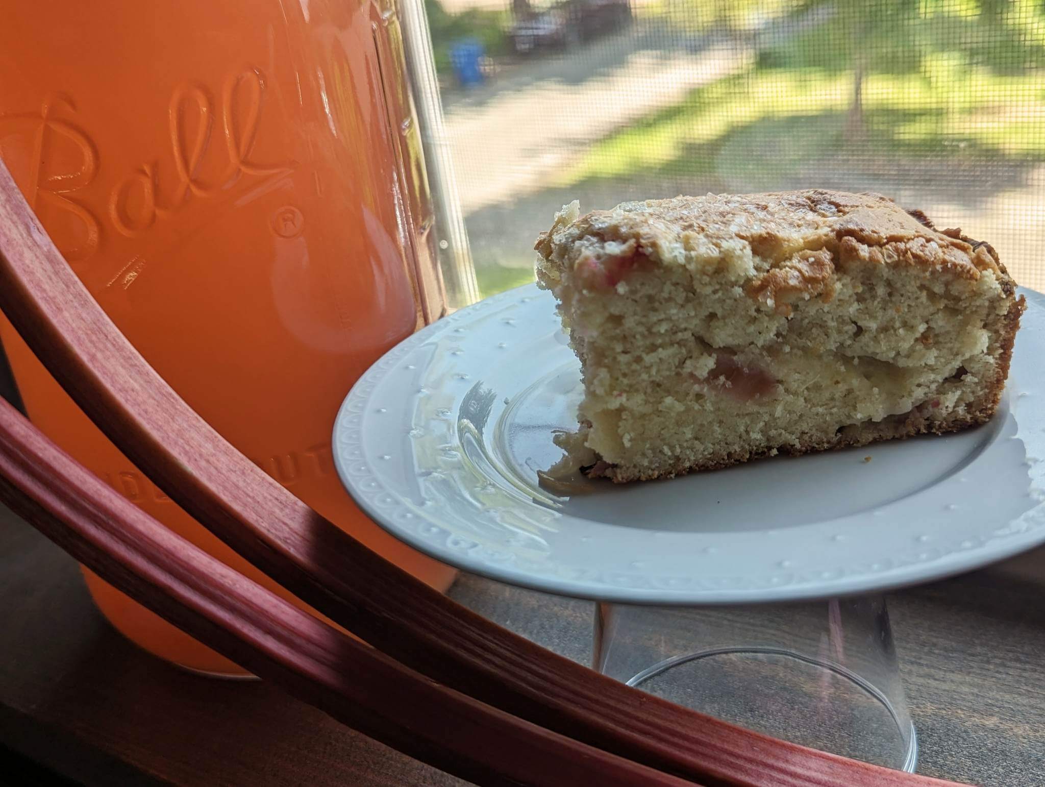 A slice of cake on a white plate next to a jar of orange drink with a window view in the background.
