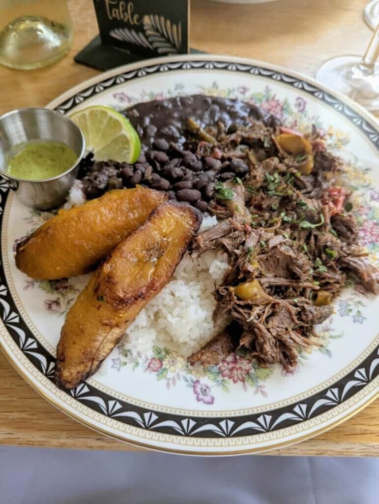 A plate of savory shredded beef, black beans, white rice, fried plantains, and a lime wedge, accompanied by a small bowl of green sauce.