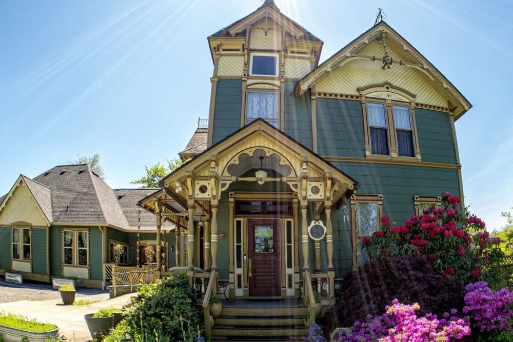 Victorian-style green house with ornate details and vibrant flower bushes in front.