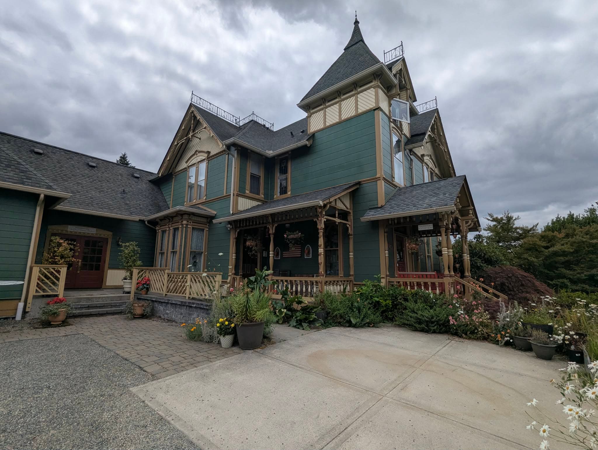 A large, ornate Victorian-style house with a green exterior and intricate architectural details, surrounded by flower beds and under a cloudy sky.